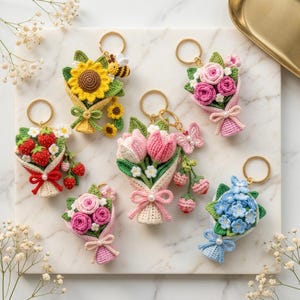 May include: A collection of six crocheted flower and fruit bouquet keychains. Designs include a sunflower with a bee, pink roses, red strawberries, pink tulips with a butterfly, small pink roses, and blue hydrangeas, all with gold rings.