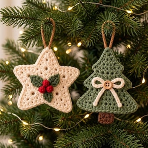 May include: Two crochet ornaments hanging from brown loops. One is a cream-colored star featuring three red berries and two green leaves. The other is a green Christmas tree shape decorated with a cream-colored yarn bow and a small wooden button.