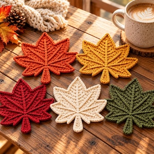 May include: Five crochet coasters shaped like maple leaves in red, orange, yellow, cream, and dark green. Each coaster features a detailed stitched vein pattern and a small stem at the base. The coasters are arranged on a wooden surface.