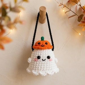 May include: A white crocheted ghost ornament featuring a small orange pumpkin hat with a green stem. The ghost has black bead eyes, a stitched smile, and pink cheeks. A black braided cord is attached to the top for hanging on a wooden hook.