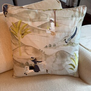 Può includere: Un cuscino quadrato con una scena paesaggistica ricamata in toni tenui di beige e verde. Il design raffigura una persona seduta a terra, un pavone e una persona a cavallo di un cavallo bianco, accentuato da motivi di palme gialle.