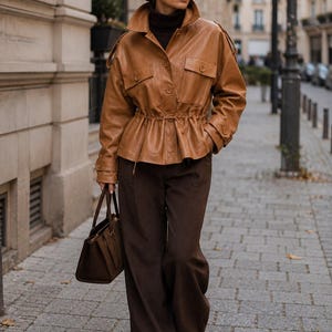 Puede incluir: Una chaqueta de cuero color canela con cintura ajustable, dos bolsillos en el pecho y puños abotonados, sobre un suéter de cuello alto marrón oscuro y pantalones anchos marrón oscuro, con un bolso de mano marrón oscuro y gafas de sol.