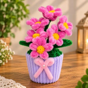 Pode incluir: Uma planta decorativa em vaso feita de limpadores de cachimbo felpudos. As flores apresentam pétalas rosa brilhante com centros amarelos, sustentadas por caules e folhas verde-escuras. O vaso é lilás com textura canelada e um laço rosa.