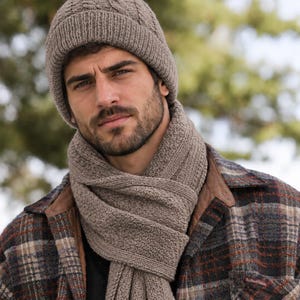 May include: A taupe knit beanie with a cable knit pattern and a matching taupe knit scarf featuring a textured stitch. The beanie has a wide folded cuff, and the scarf is wrapped around the neck, worn over a brown and grey plaid flannel shirt.