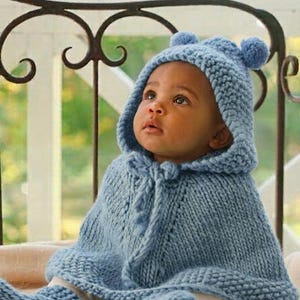 May include: A light blue knitted poncho with a hood featuring two small pom-poms on top. The garment is crafted from thick yarn with a textured stitch pattern and includes a front tie closure with matching knitted tassels at the neckline.