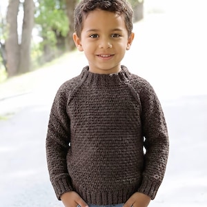 Könnte beinhalten: Ein dunkelbrauner Langarmpullover mit strukturiertem Strickmuster, geripptem Rundhalsausschnitt sowie gerippten Bündchen an Ärmeln und Taille. Der Pullover wird mit einer hellblauen Jeans kombiniert, was ein schlichtes und funktionales Outfit ergibt.