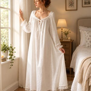 May include: A floor-length white nightgown featuring long sleeves with ruffled cuffs, a square neckline with a delicate tie closure, and a wide lace trim along the bottom hem. The garment is made of a lightweight fabric with a soft, flowing silhouette.