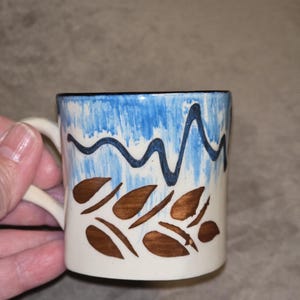 May include: A ceramic mug with a cream-colored base featuring a hand-painted design. The upper portion has a blue brushed wash with a dark blue wavy line, while the lower section displays a pattern of brown leaf-like shapes on the cream surface.