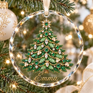 May include: A circular clear glass Christmas ornament featuring a detailed illustration of a green pine tree decorated with emerald green crystals, white pearls, and gold chains. The name Emma is written in gold script at the bottom.