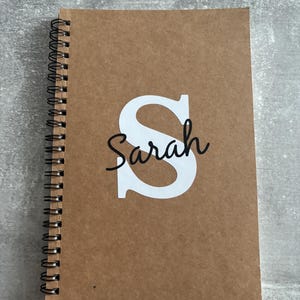 May include: A spiral-bound notebook with a brown kraft paper cover. The front cover features a large white capital letter S with the name Sarah written in black cursive script layered over the centre of the letter.