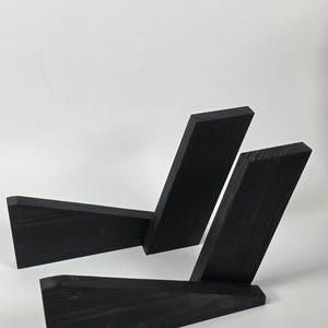 May include: A black wooden stand composed of two interlocking geometric pieces. Each piece features a long, angled base and a vertical support, creating a V-shaped profile. The wood grain is visible through the matte black finish on all surfaces.