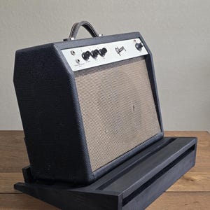 May include: A vintage Gibson guitar amplifier featuring a black textured exterior, a silver control panel with black knobs, and a tan fabric speaker grille. The amplifier is positioned on a black wooden angled stand with a horizontal slot design.