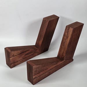 May include: Two L-shaped wooden supports crafted from dark brown stained timber with a visible wood grain texture. Each piece features a horizontal base and an angled upright section joined together, creating a sturdy, angular structural component.