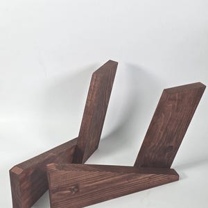 Puede incluir: Dos tablas de madera teñidas de marrón oscuro con un patrón de veta visible, cada una cortada en ángulo para formar una estructura de soporte inclinada. Las piezas están colocadas para crear un marco abierto y angular con bordes planos.