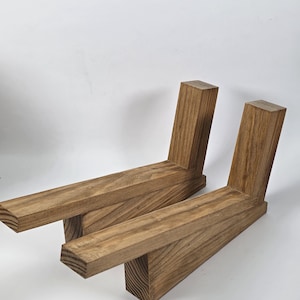 May include: Two L-shaped wooden furniture legs featuring a natural brown wood grain finish. Each piece consists of a long horizontal base section joined to a shorter, angled vertical support post with visible wood texture and grain patterns.