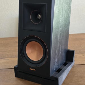 May include: A black bookshelf speaker with a wood grain finish, featuring a copper-colored woofer cone and a horn-loaded tweeter. The front panel displays the Klipsch brand name in copper lettering. The speaker is mounted on a black wooden stand.