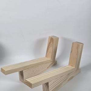 May include: Two L-shaped wooden stands crafted from light-colored natural wood with visible grain patterns. Each stand features a horizontal base and a vertical support piece joined together, designed to hold items in an upright position.