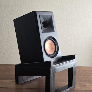 Puede incluir: Un altavoz de estantería negro Klipsch con un woofer de color cobre y un tweeter de bocina cuadrado, apoyado sobre un soporte de madera negro inclinado. El altavoz presenta una textura de veta de madera y el logotipo de Klipsch es visible.