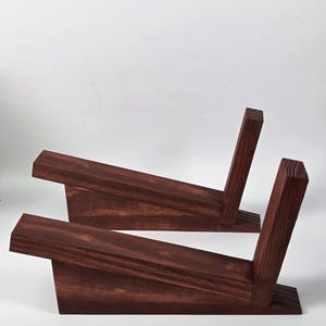 May include: Two dark brown wooden stands, each featuring a long, angled horizontal support piece attached to a vertical back support. The wood grain is visible throughout the structure, showcasing a deep, rich, reddish-brown stained finish.