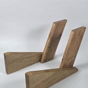 Puede incluir: Dos soportes de madera a juego, cada uno construido con dos piezas anguladas de madera marrón claro natural con vetas visibles. Los soportes presentan un diseño geométrico de perfil bajo con una base plana y un brazo de soporte inclinado.