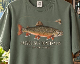 Puede incluir: Una camiseta de manga corta color verde salvia con una ilustración detallada de una trucha de arroyo, acompañada de una mosca de pesca y un sedal. El texto SALVELINUS FONTINALIS y Brook Trout está impreso en blanco bajo el gráfico del pez.