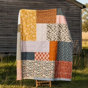 May include: A patchwork quilt featuring a variety of rectangular fabric blocks in mustard yellow, burnt orange, navy blue, white, and soft pink. The design includes floral, geometric, and solid patterns with visible grid-like quilting stitches.