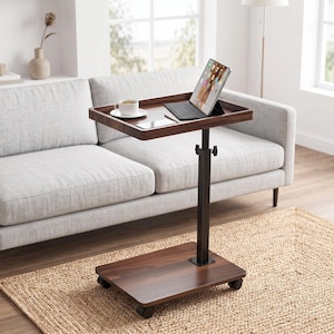 May include: A dark brown wooden rolling side table with a rectangular top and a height-adjustable black metal central column. The base features four small wheels for mobility. The table surface holds a white coffee cup, a smartphone, and a tablet.