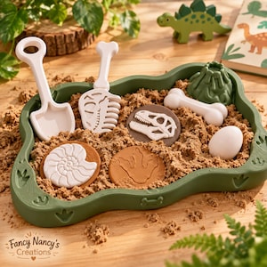May include: A green sensory tray filled with sand containing white plastic dinosaur-themed tools, including a shovel and a bone-shaped rake, along with circular fossil stamps, a white egg, a bone, and a small green volcano figure.