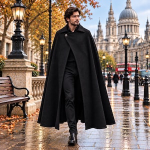 May include: A long black wool cape featuring a single metallic button closure at the collar, worn over a black shirt and black trousers. The garment drapes to the mid-calf, providing a structured silhouette with a wide, flowing hemline.