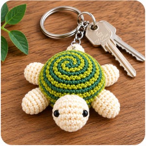 Peut inclure: Un porte-clés tortue en crochet avec une tête et quatre membres de couleur crème, doté d'une carapace présentant un motif en spirale alternant des nuances de vert foncé et clair. La tortue est fixée à un anneau en métal argenté avec deux clés.