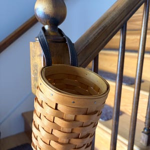 Peut inclure: Un panier rond en bois tressé avec une finition brun clair et une sangle en cuir marron foncé fixée au bord. Le panier présente une construction en éclisses tressées avec de petits rivets métalliques fixant le bord et la sangle au corps.