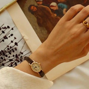 May include: A gold-toned oval wristwatch with a dark brown textured leather strap and a white dial featuring black Roman numerals. The watch is worn on the wrist alongside a gold-toned ring with a small brown stone, set against a blurred background.