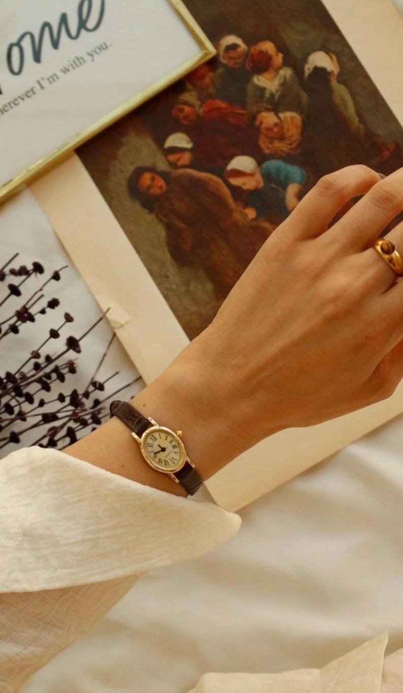 May include: A gold-toned oval wristwatch with a dark brown textured leather strap and a white dial featuring black Roman numerals. The watch is worn on the wrist alongside a gold-toned ring with a small brown stone, set against a blurred background.