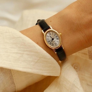 May include: A gold-toned oval wristwatch featuring a white dial with black Roman numeral hour markers and black hands. The watch includes the word QUARTZ printed on the face and is secured with a slim, smooth black leather strap with a gold buckle.