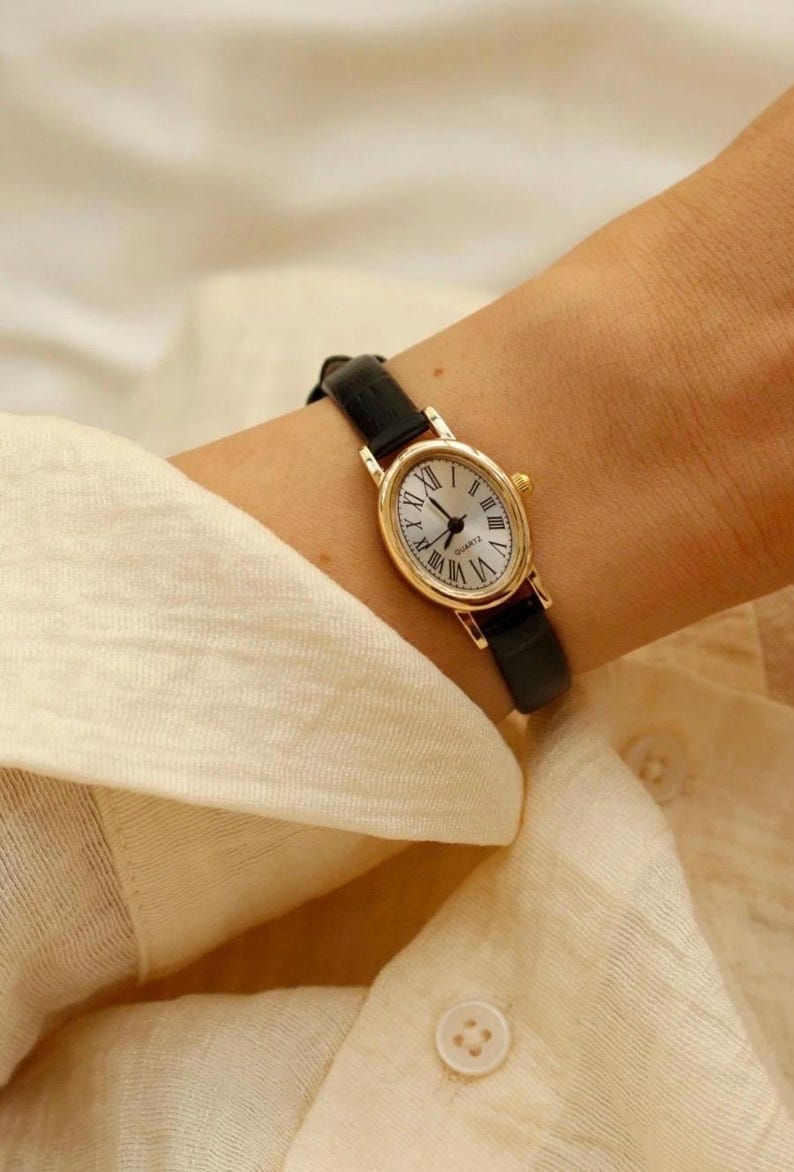 May include: A gold-toned oval wristwatch featuring a white dial with black Roman numeral hour markers and black hands. The watch includes the word QUARTZ printed on the face and is secured with a slim, smooth black leather strap with a gold buckle.