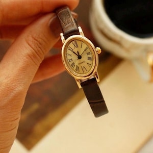 May include: An oval-shaped gold-tone quartz watch featuring a cream-colored dial with black Roman numeral hour markers and thin black hands. The timepiece is attached to a slim, dark brown textured leather strap with a crocodile-like pattern.