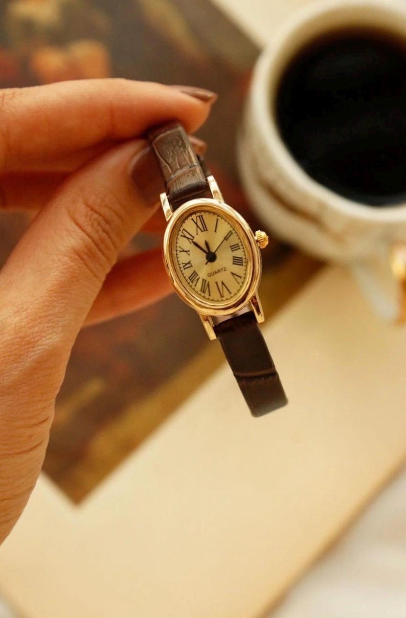 May include: An oval-shaped gold-tone quartz watch featuring a cream-colored dial with black Roman numeral hour markers and thin black hands. The timepiece is attached to a slim, dark brown textured leather strap with a crocodile-like pattern.