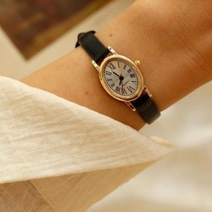 May include: An oval-shaped wristwatch with a gold-tone metal case and a black leather strap. The watch face features a white background with black Roman numeral hour markers and black hands. The word QUARTZ is printed in small black letters on the dial.