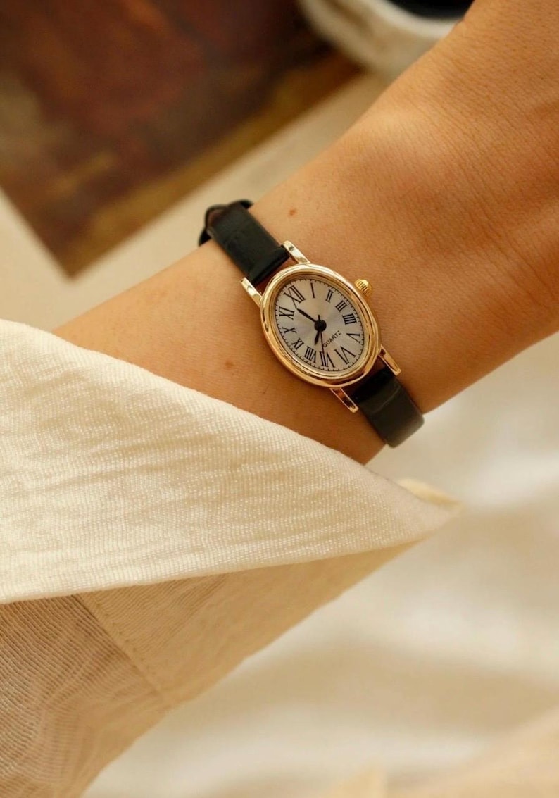 May include: An oval-shaped wristwatch with a gold-tone metal case and a black leather strap. The watch face features a white background with black Roman numeral hour markers and black hands. The word QUARTZ is printed in small black letters on the dial.