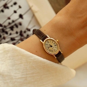 May include: An oval-shaped gold-tone wristwatch featuring a cream-colored dial with black Roman numeral hour markers and thin black hands. The watch is equipped with a dark brown textured leather strap and a small gold-tone winding crown on the side.