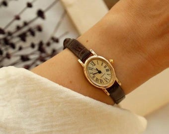 May include: An oval-shaped gold-tone wristwatch featuring a cream-coloured dial with black Roman numeral hour markers and thin black hands. The watch is equipped with a dark brown textured leather strap and a small gold-tone winding crown on the side.