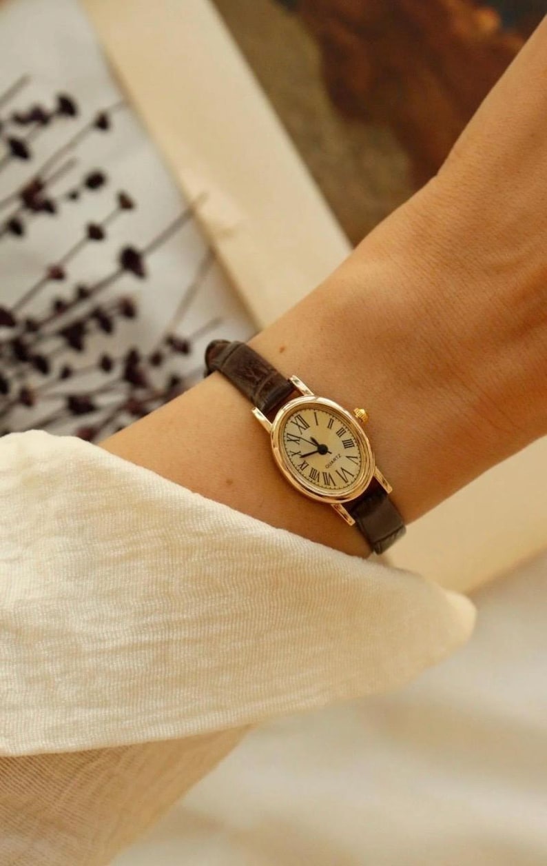 May include: An oval-shaped gold-tone wristwatch featuring a cream-colored dial with black Roman numeral hour markers and thin black hands. The watch is equipped with a dark brown textured leather strap and a small gold-tone winding crown on the side.