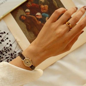 May include: A gold-toned oval wristwatch with a dark brown textured leather strap and a white dial featuring black Roman numerals. A gold-toned ring with a small, round, brown stone is worn on the ring finger. The watch has a classic, slim design.
