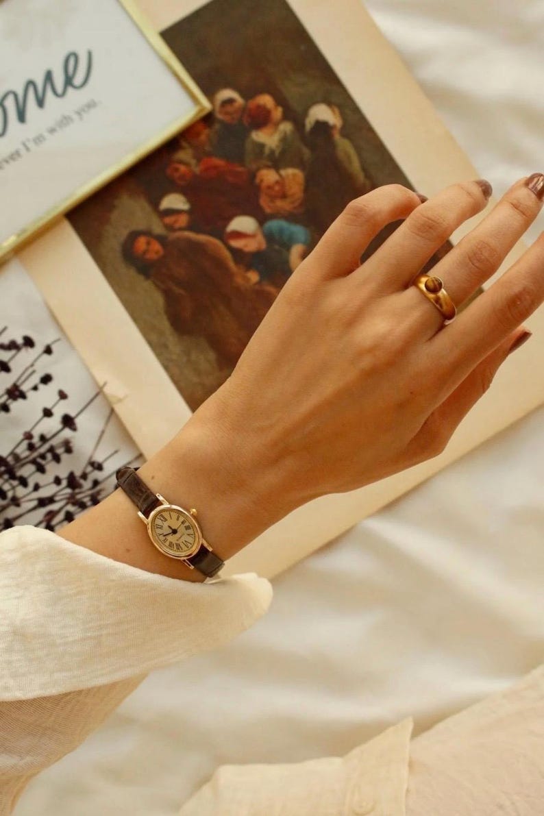May include: A gold-toned oval wristwatch with a dark brown textured leather strap and a white dial featuring black Roman numerals. A gold-toned ring with a small, round, brown stone is worn on the ring finger. The watch has a classic, slim design.