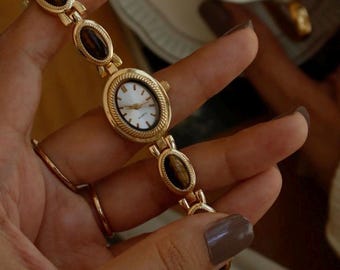 May include: A gold-tone analog wristwatch featuring an oval-shaped white face with black markers and hands. The watch band is composed of alternating oval links, each set with a smooth, brown tiger's eye gemstone, secured by a metal clasp.