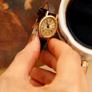 May include: An oval-shaped gold-tone wristwatch with a cream-colored dial, black Roman numeral hour markers, and the word "QUARTZ" printed on the face. The watch features a dark brown leather strap with a textured finish and a gold-tone buckle.