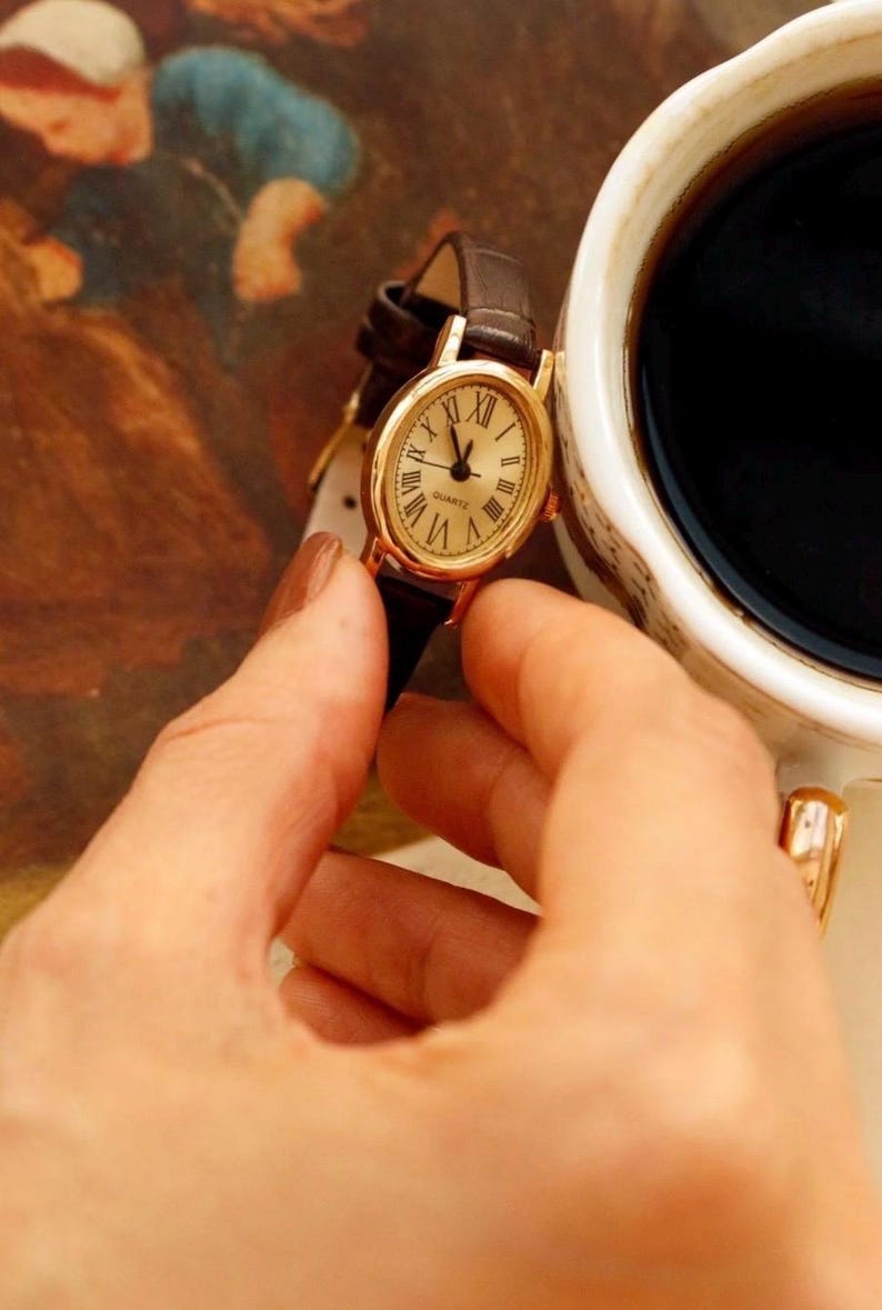 May include: An oval-shaped gold-tone wristwatch with a cream-colored dial, black Roman numeral hour markers, and the word "QUARTZ" printed on the face. The watch features a dark brown leather strap with a textured finish and a gold-tone buckle.