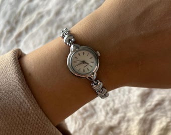 May include: A silver-tone analog quartz wristwatch featuring a round white dial with simple silver markers and hands. The watch has a delicate link bracelet composed of small, heart-shaped metal segments that connect to the circular watch case.