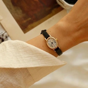 May include: A gold-toned oval wristwatch with a white face, black Roman numeral hour markers, and thin black hands. The watch features a slim black leather strap with a gold-toned buckle, secured around a wrist with a light-colored long-sleeved shirt.