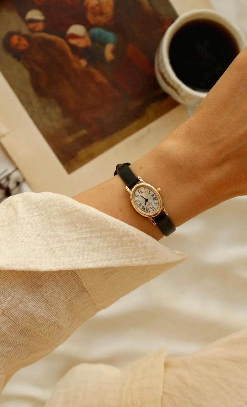 May include: A gold-toned oval wristwatch with a white face, black Roman numeral hour markers, and thin black hands. The watch features a slim black leather strap with a gold-toned buckle, secured around a wrist with a light-colored long-sleeved shirt.